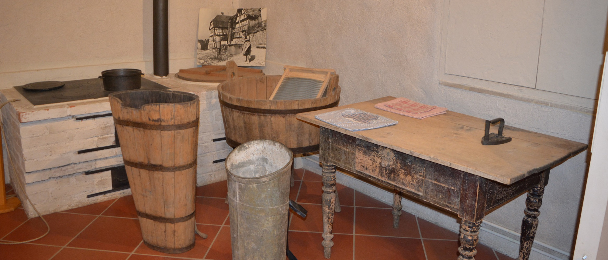 Blick in einen Ausstellungsraum im Volkskundemuseum. Rechts steht ein großer Tisch mit einem alten Glätteisen. Links daneben stehen Waschzuber und ein Herd mit Waschkessel.