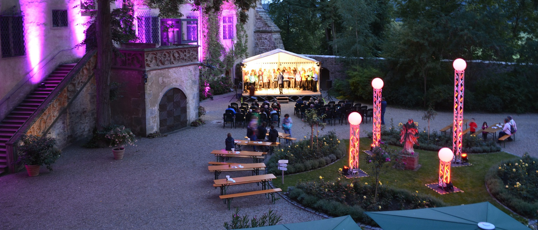 Schlossinnenhof in abendlicher Stimmung. Der Schlossinnenhof ist mit Leuchten geschmückt. Sie leuchten lila und rosa. Auf einer Bühne im Hintergrund stehen Musikerinnen und Musiker. Auf Biertischgarnituren sitzen Gäste.