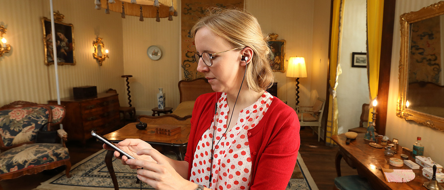 Eine Frau steht in einem Zimmer des Schlosses. Sie hält ein Smartphone in der Hand und hat Kopfhörer im Ohr.