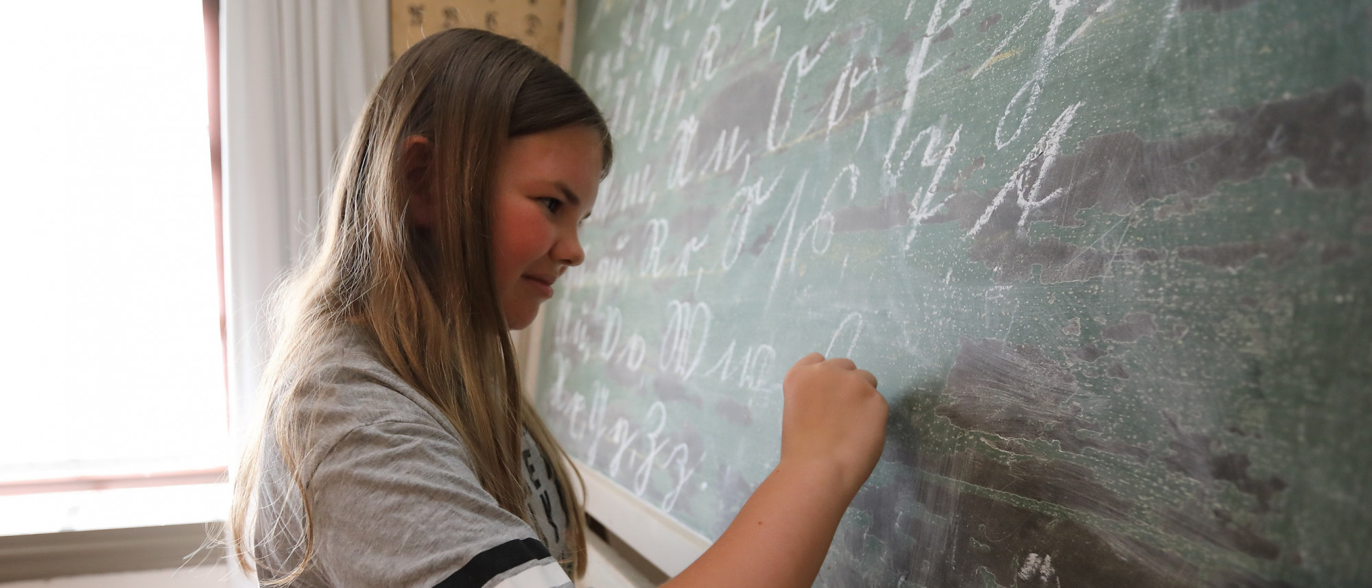 Ein Mädchen steht an einer alten Schiefertafel und schreibt auf diese mit Kreide.