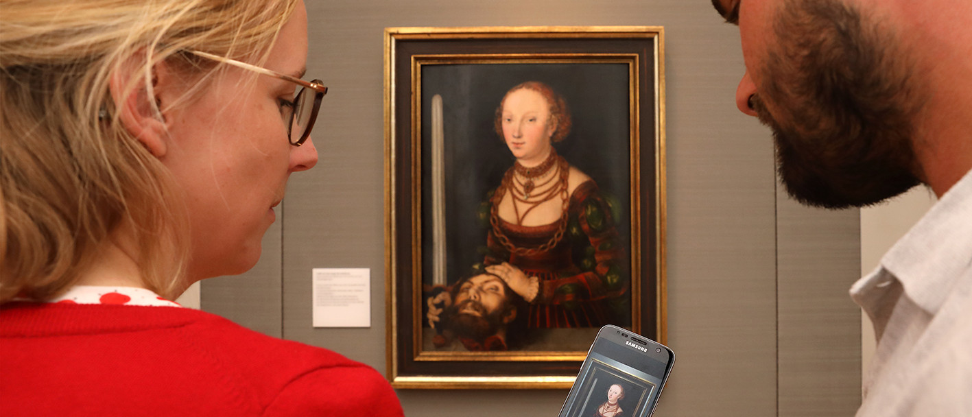 An einer Wand hängt ein Gemälde: Judith mit dem Haupt des Holofernes von Lucas Cranach dem Älteren. Eine Frau und ein Mann stehen vor dem Gemälde und schauen auf ein Smartphone. Auf dem Smartphone ist das gleiche Gemälde zu sehen.