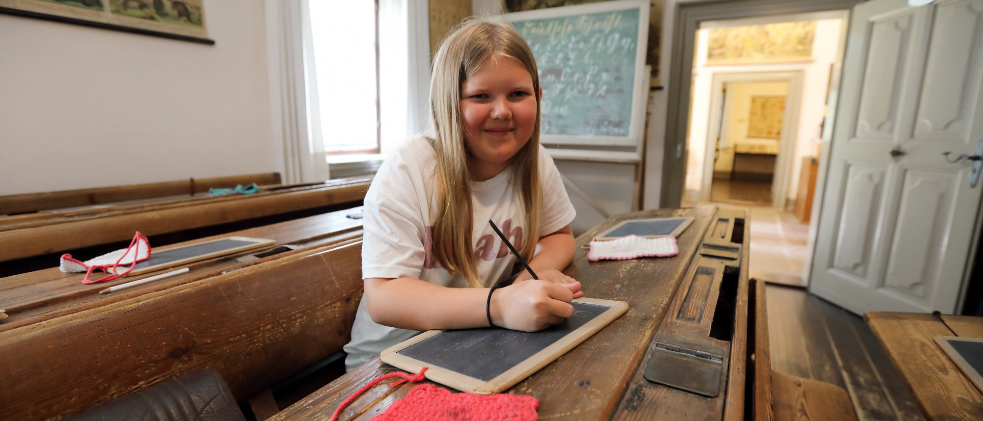 Ein Mädchen sitzt auf einer Schulbank in einem alten Klassenzimmer. Sie schreibt mit einem Griffel auf eine Schiefertafel.