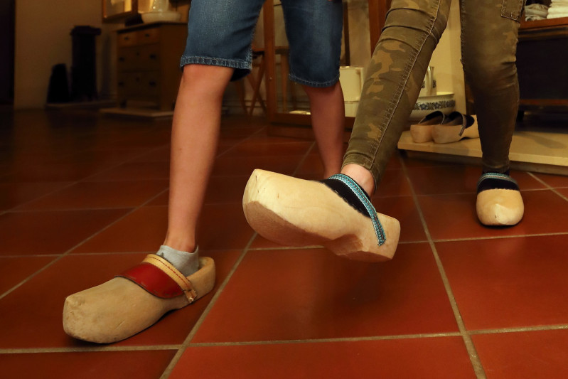 Die Füße von zwei Kindern stecken in Holzpantoffeln. Mit diesen Schuhen laufen sie über einen dunkelroten Fließenboden. 