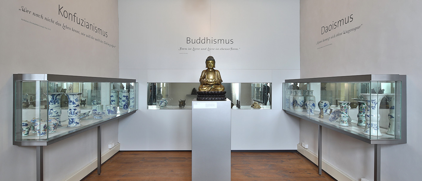 Blick in einen Ausstellungsraum der ostasiatischen Kunst. In der Mitte steht auf einem grauen Sockel eine Buddha-Skulptur. Links, dahinter und rechts an den Wänden befinden sich Vitrinen mit Vasen, Tellern und Skulpturen.