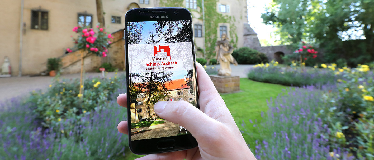 Eine Hand hält im Aschacher Schlosspark ein Smartphone. Auf dem Smartphone ist die Startseite der App &quot;Graf-Luxburg-Museum&quot; zu sehen.