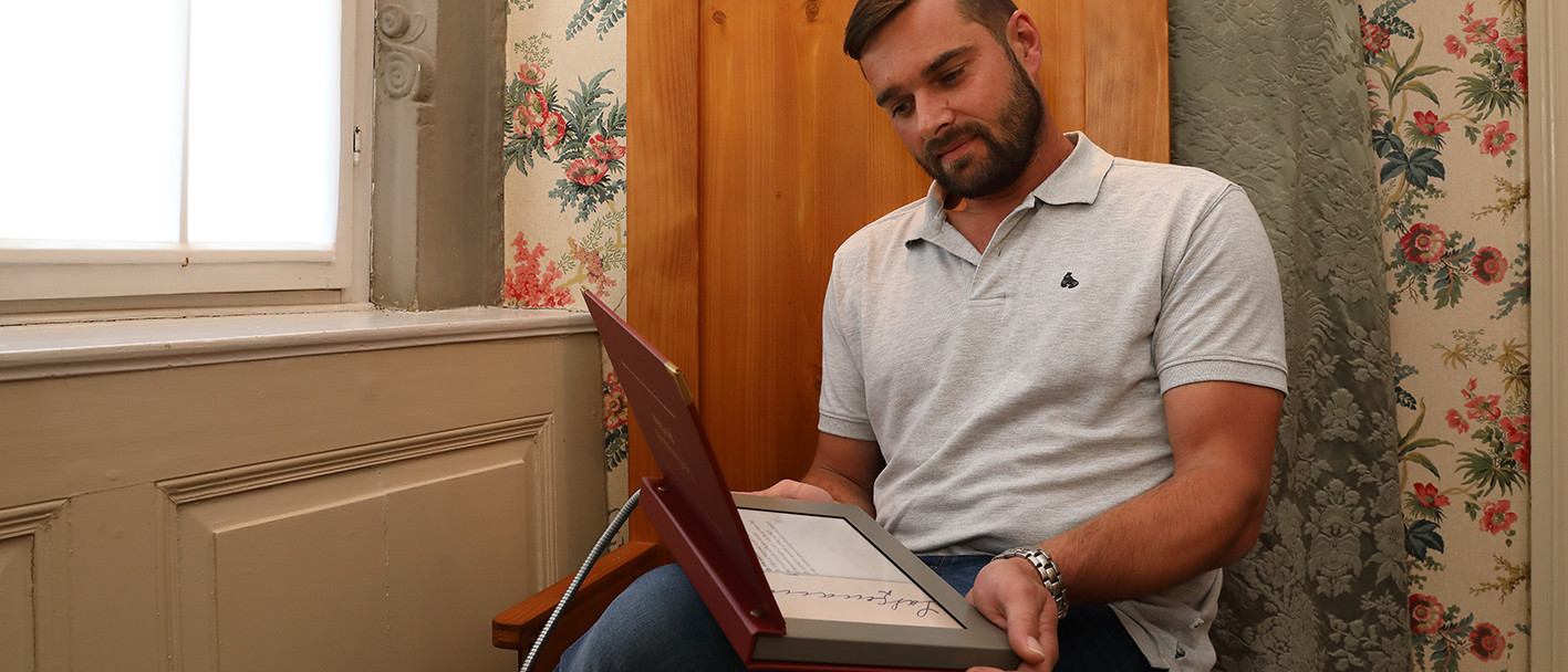 Ein Mann sitzt auf einer braunen Bank mit hoher Rückenlehne und hält ein Tablet in der Hand. Im Hintergrund ist die geblümte Tapete des Schlosssalons zu sehen.