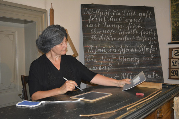 Ein Frau mit schwarzem Kleid sitzt am Lehrerpult. Die Lehrerin trägt eine altmodische Perücke. Die Lehrerin korrigiert gerade. Die Tafel neben ihr ist in alter Schrift beschrieben.