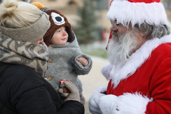 Ein kleines Kind auf dem Arm seiner Mama bekommt vom Weihnachtsmann eine Süßigkeit überreicht.